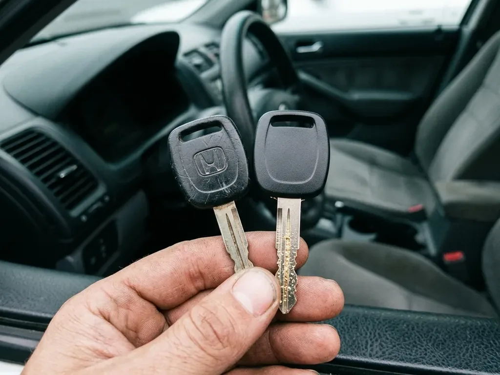 Hand holding a worn original chip key and a fresh cut clone side by side with an older Honda dashboard behind
